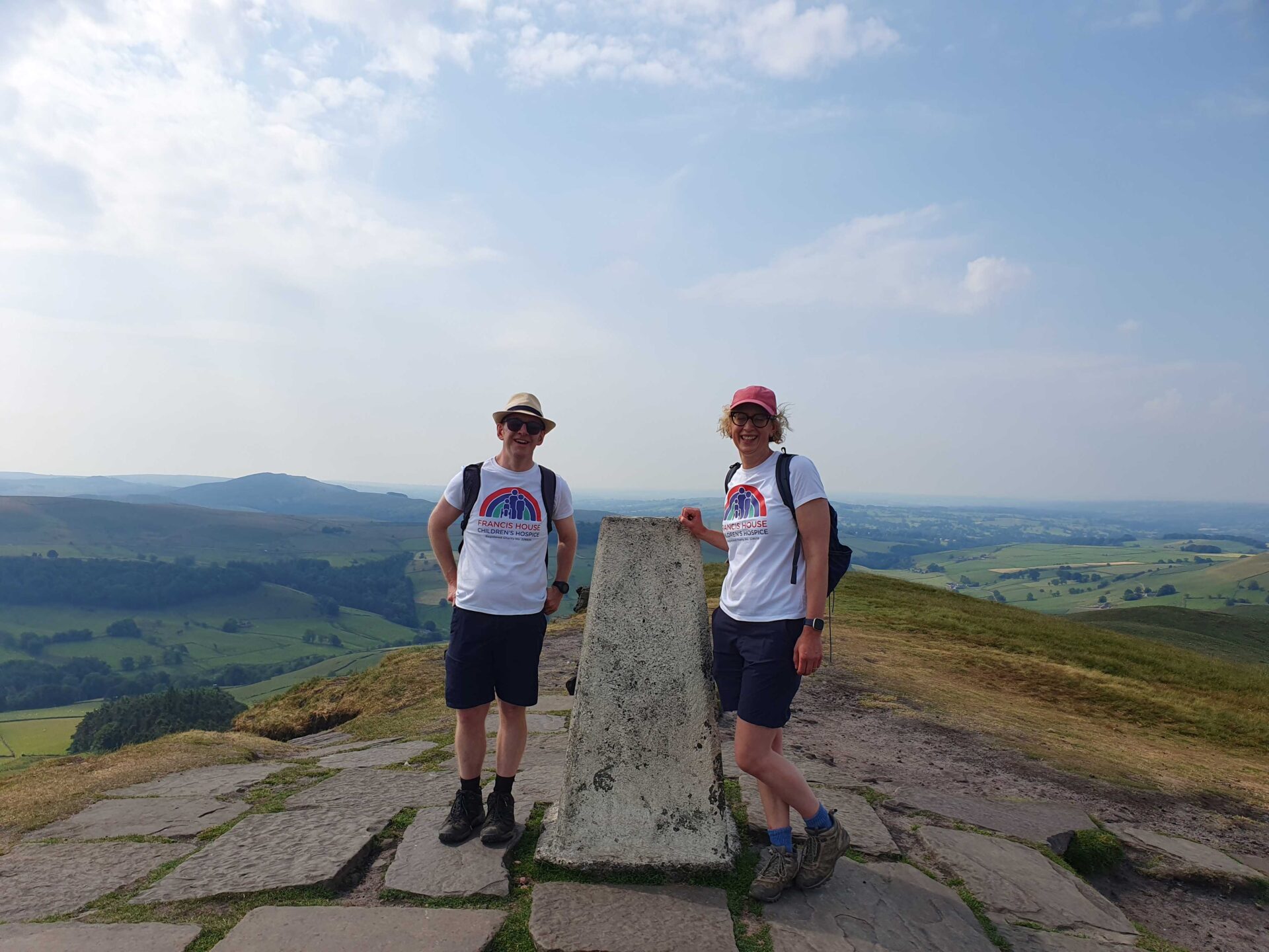 Two walkers stand eithe side of Shutlingsloe's trig point