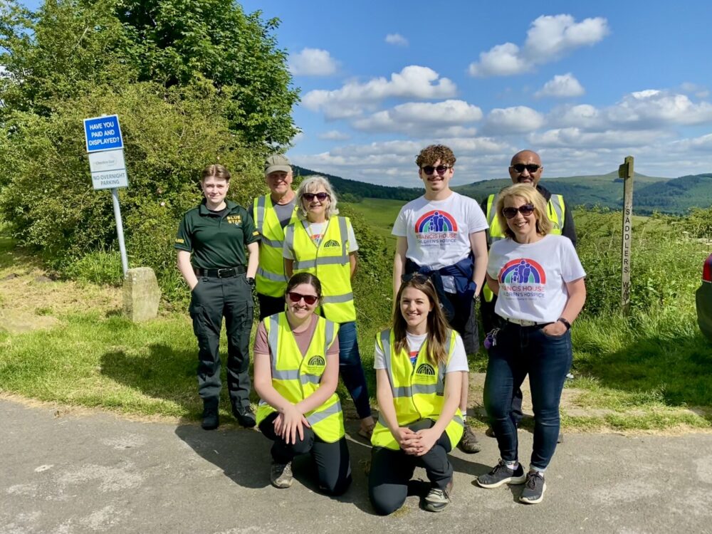 8 people (5 wearing Francis House hi-vis jackets, two in Francis House T-shirts and one in a medical uniform) smile to the camera infront of the countryside landscape on a sunny day