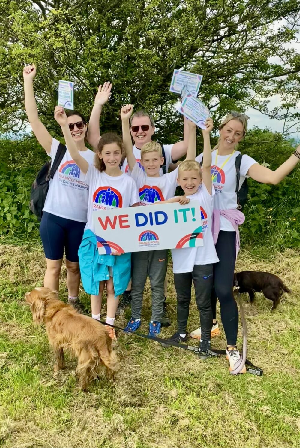 Three adults and three children hold a board saying 'WE DID IT' and cheer to the camera