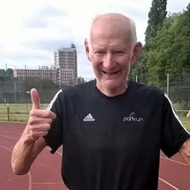Elderly man wearing a black top with his thumbs up stood on a running track.