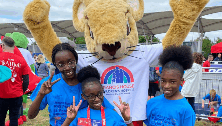 Three young girls wearing Great Manchester Junior Run T-shirts pose for the camera with Francis Mouse, the giant furry mouse mascot, by the running track