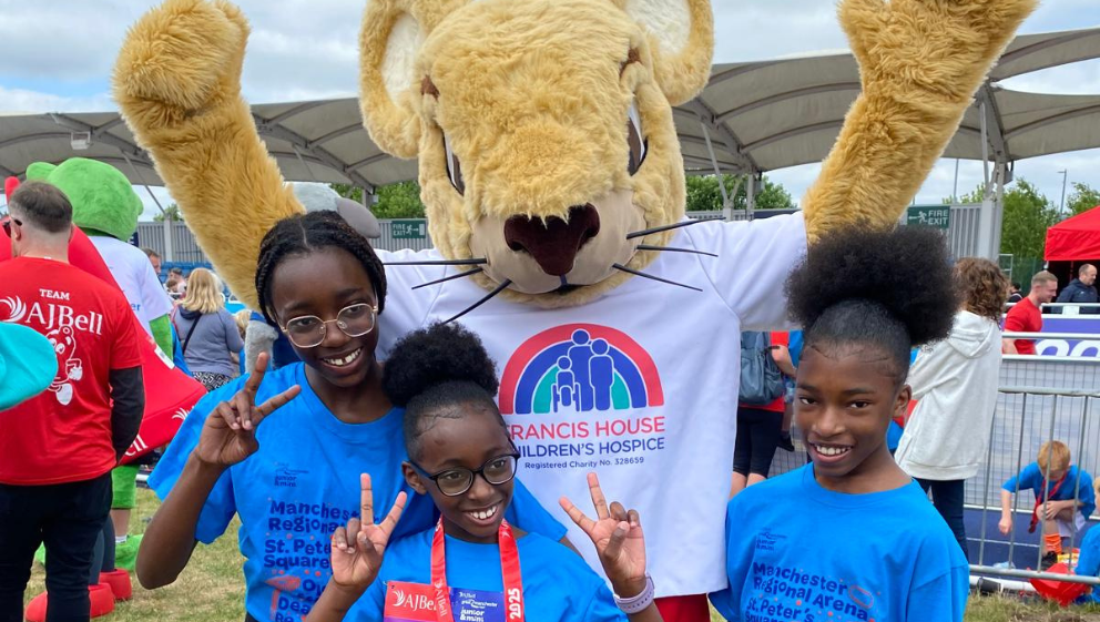 Three young girls wearing Great Manchester Junior Run T-shirts pose for the camera with Francis Mouse, the giant furry mouse mascot, by the running track