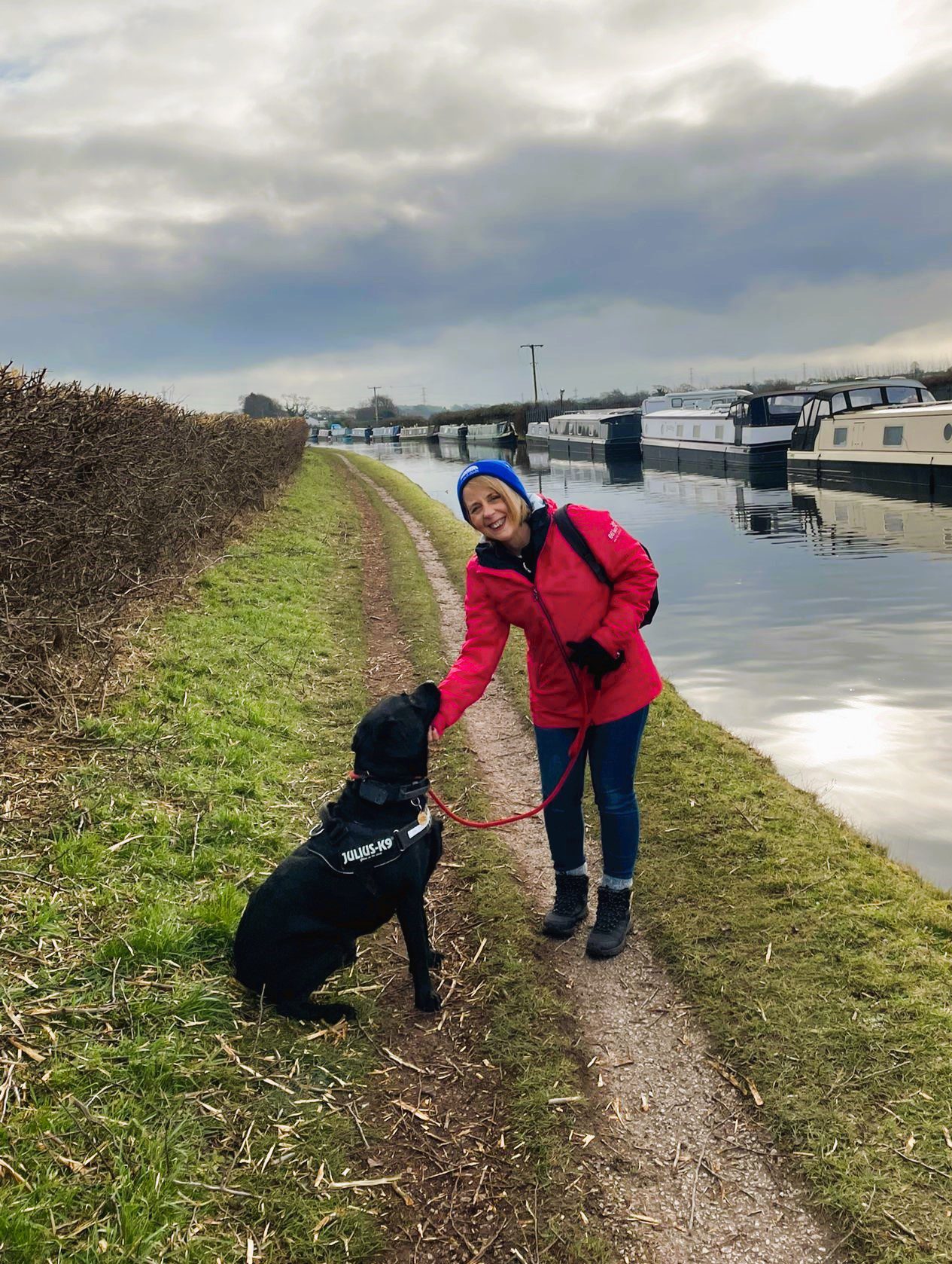 A lady wearing a Francis House beanie poses for a photo with a black Labrador by the side of the canal along the Lymm Loop.