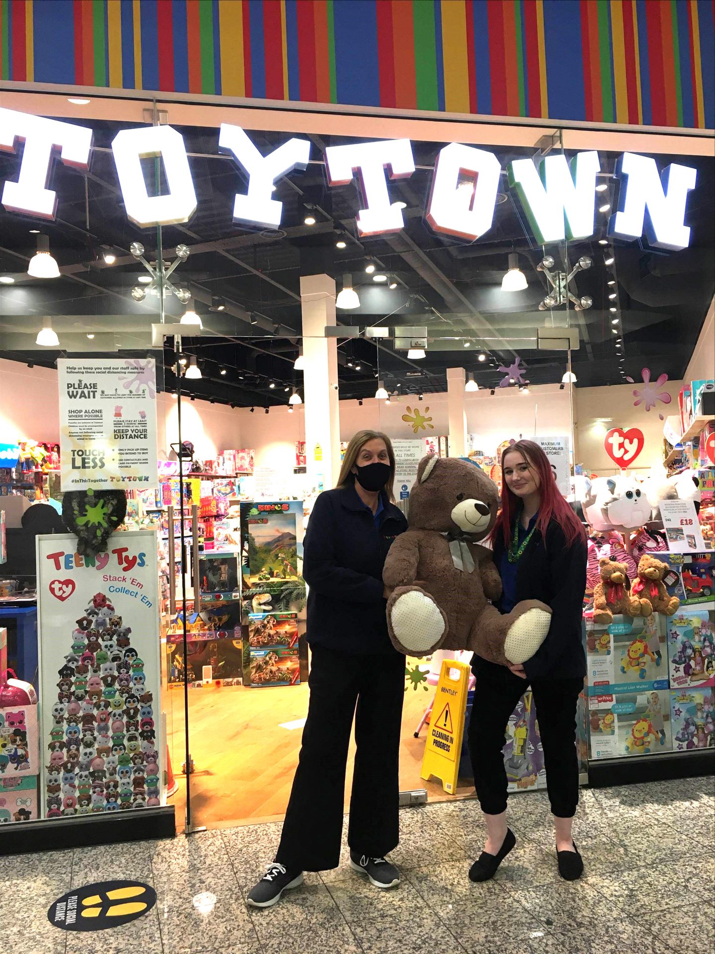 Two women holding a teddy bear outside a toy shop