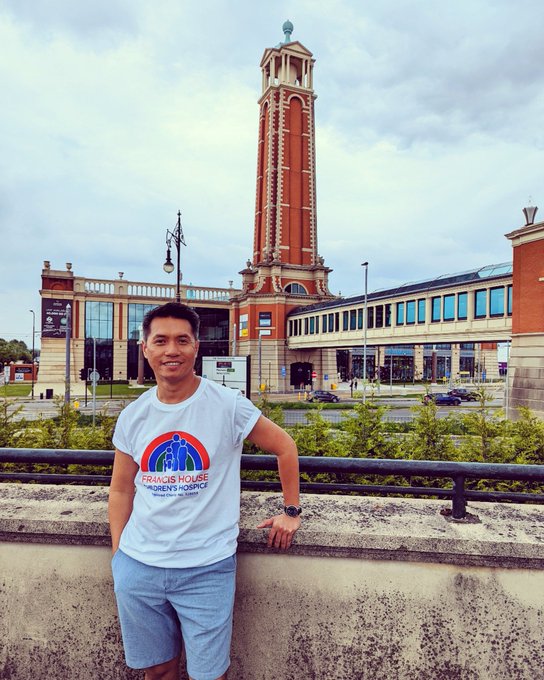 A man in a Francis House T-shirt stands in front of a wall with the Trafford Palazzo tower seen in the background