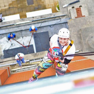 A bird's-eye view photo taken looking down from a tall tower (Trafford Palazzo) where a woman wearing a Francis House t-shirt and a helmet is making her descent and looking up to smile at the camera