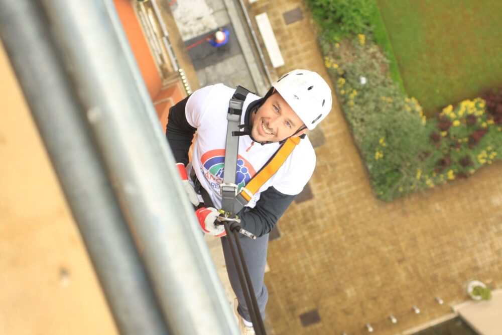 A bird's-eye view photo taken looking down from a tall tower (Trafford Palazzo) where a man wearing a Francis House t-shirt and a helmet is making his descent and looking up to smile at the camera