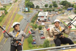 Two smiling women hang backwards off the edge of the tower where the camera is positioned, with their hands in the air. The view down to the car park far below can be seen behind them.