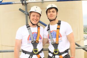 Two men wearing Francis House t-shirts, safety harnesses and helmets stand together facing the camera