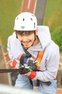 A man in a helmet, harness and a grey Francis House hooded top smiles to camera as he hangs from the rope at the top of tower where the camera is positioned, ready to descend
