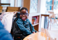 Child sitting in a wheelchair sat by a table with toys on it smiling
