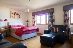Bedroom with double bed and purple duvet, brown leather sofas and colourful curtains