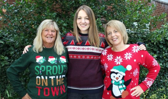 Three woman wearing Christmas jumpers stood in front of a tree
