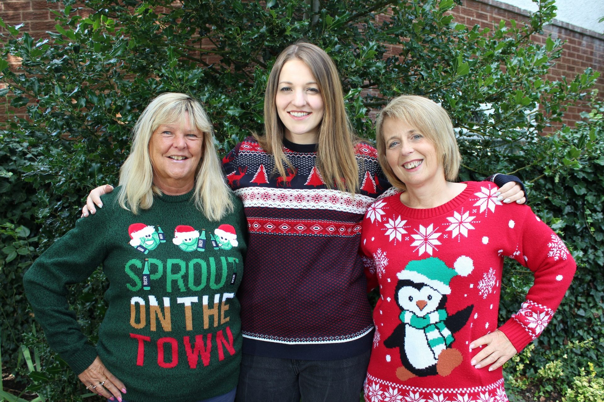 Three woman wearing Christmas jumpers stood in front of a tree