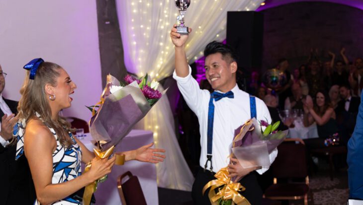 Person holding trophy in the air with other person holding flowers after winning a dance competition