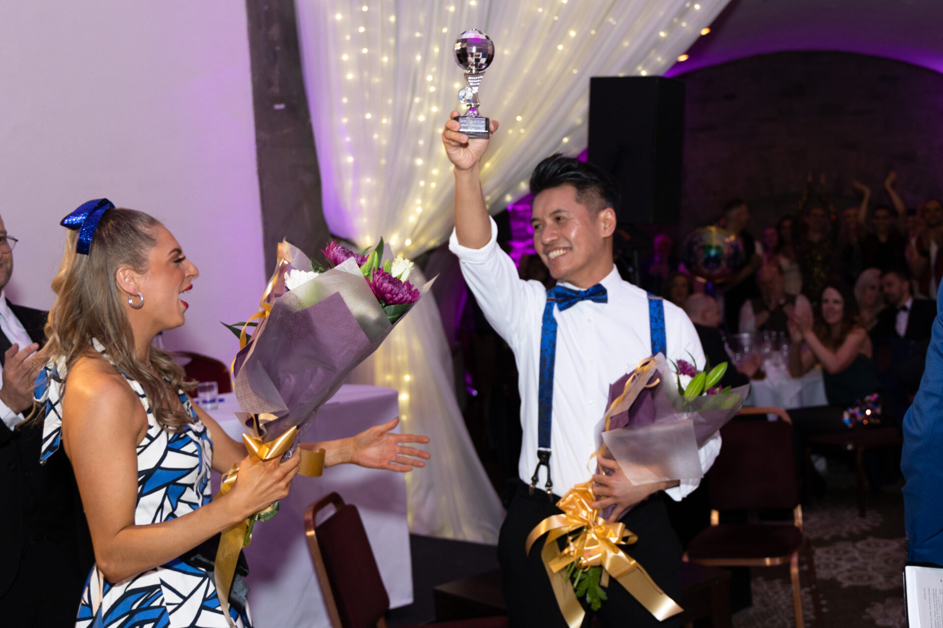 Person holding trophy in the air with other person holding flowers after winning a dance competition