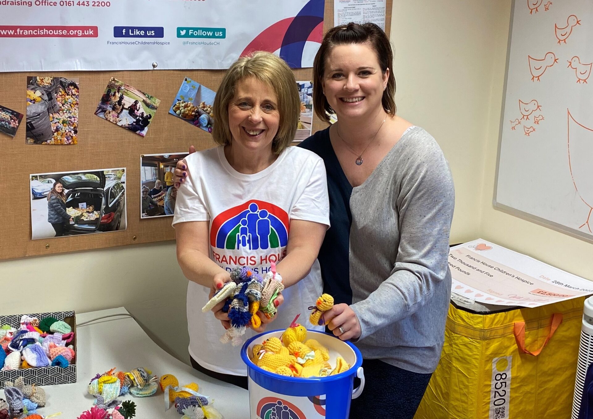 Two women holding knitted chicks stood by a bucket of knitted chicks