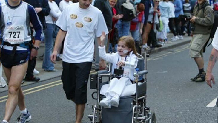 Girl in a wheelchair smiling accompanied by a runner with crowds behind