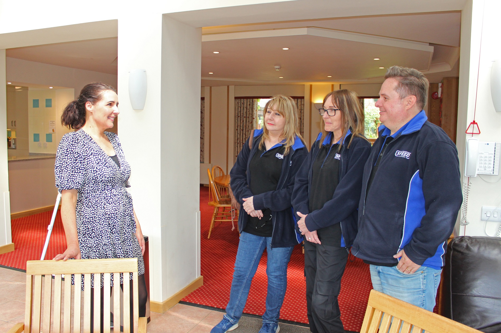 Three people in uniforms listening to a woman talking stood in a conservatory