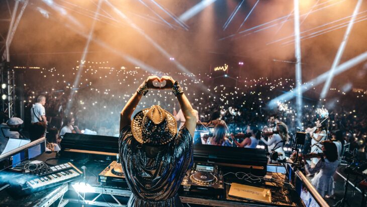 DJ on stage holding hands in a heart shape stood on stage facing a large crowd with light show.