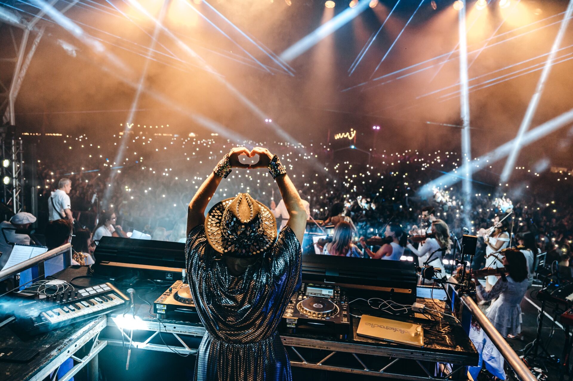 DJ on stage holding hands in a heart shape stood on stage facing a large crowd with light show.