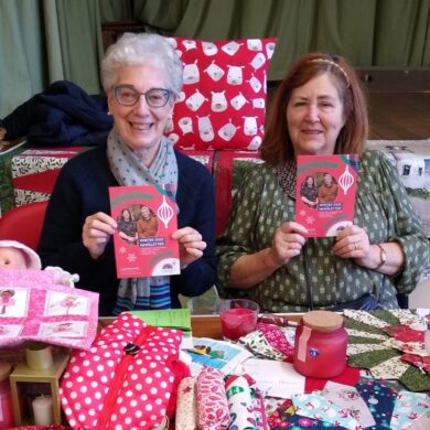 Two women holding booklets sat in front of craft items for sale on a stall in a village hall
