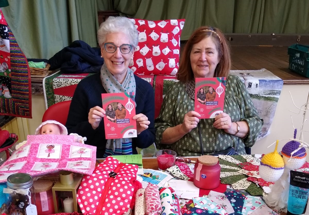 Two women holding booklets sat in front of craft items for sale on a stall in a village hall