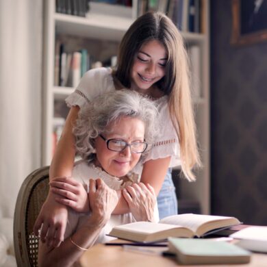 A young girl stood behind an elderly lady with her hands on her shoulders looking down at a book