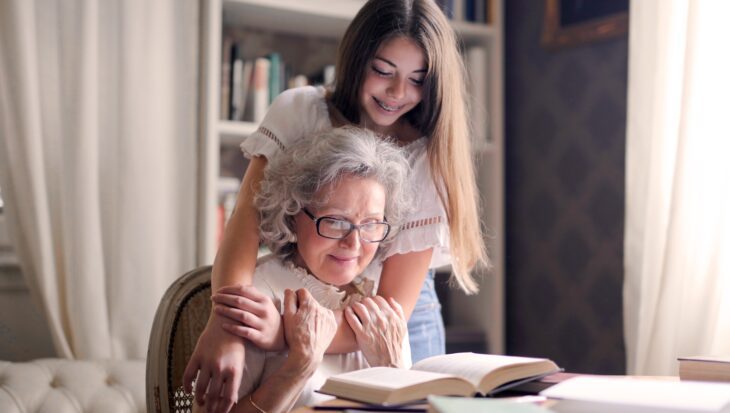A young girl stood behind an elderly lady with her hands on her shoulders looking down at a book