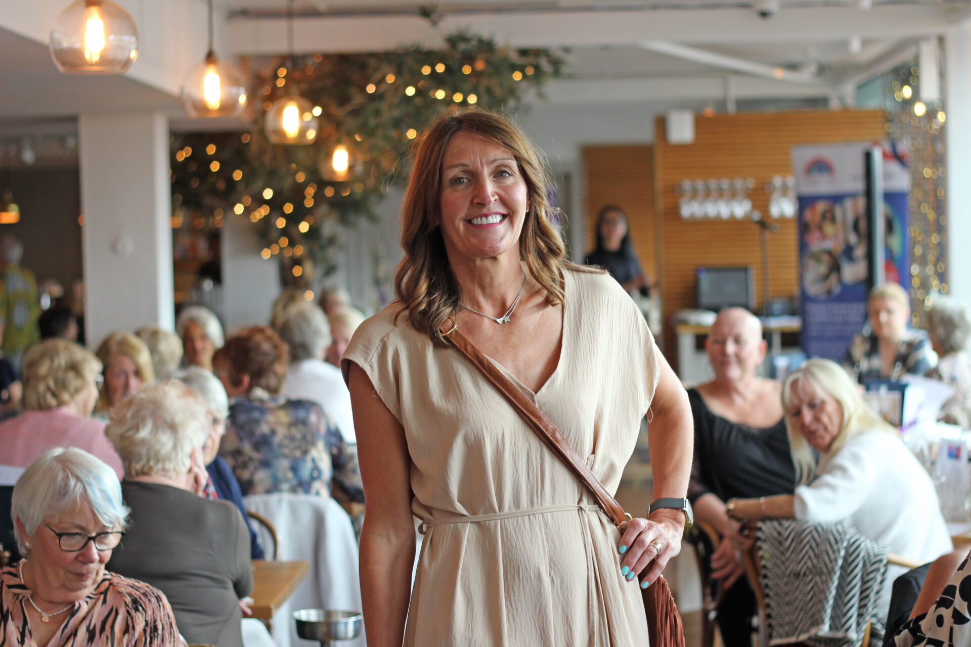 Woman wearing a beige jumpsuit holding a hand bag stood in a restaurant