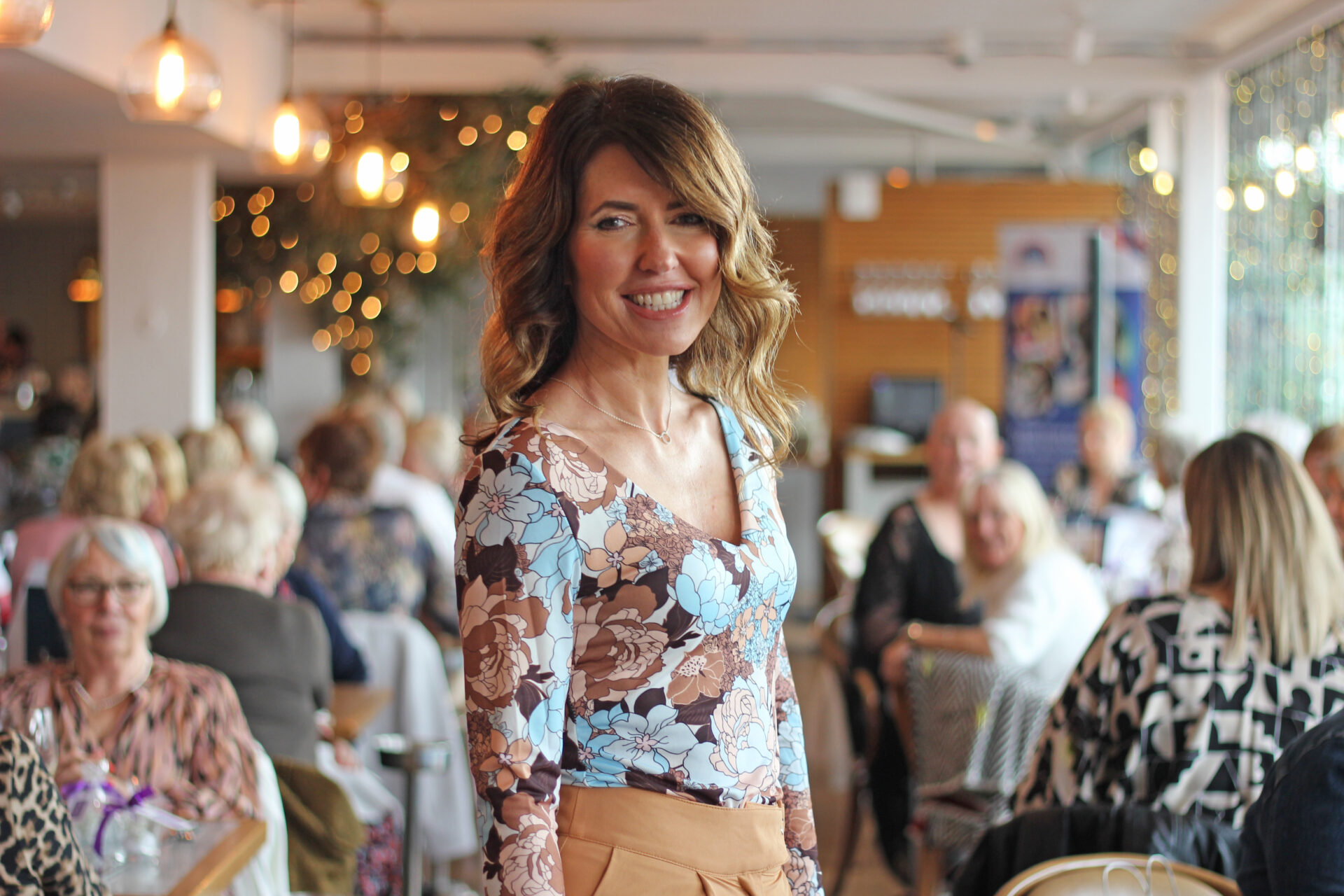 Woman wearing a multi-coloured top and tan trousers stood in a restaurant