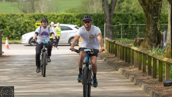 Tour de Manc riders, one wearing a Francis House t-shirt, approach the finish line on their bikes