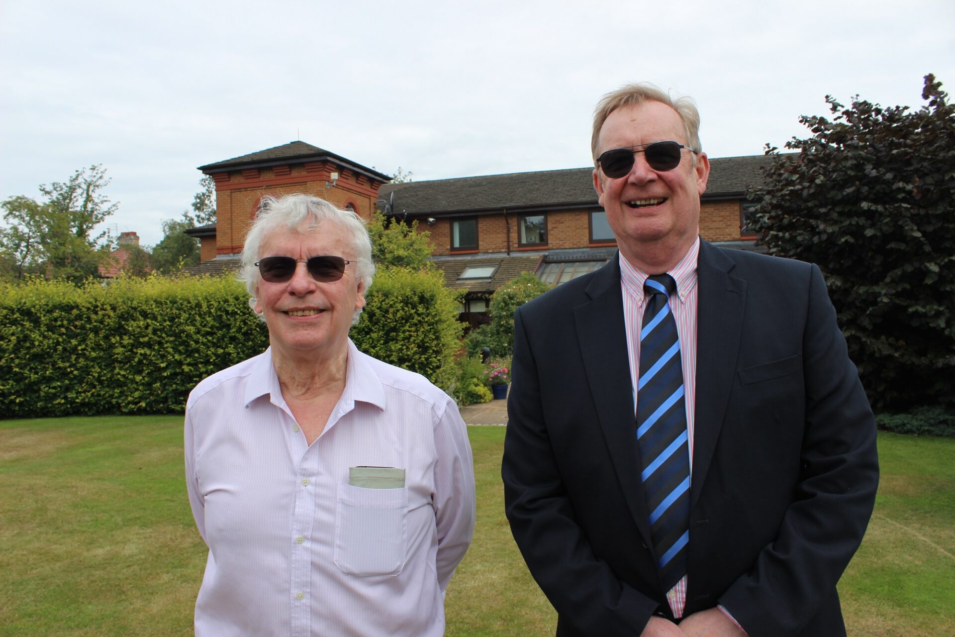 Two men wearing sunglasses stood on a lawn infront of a brick building