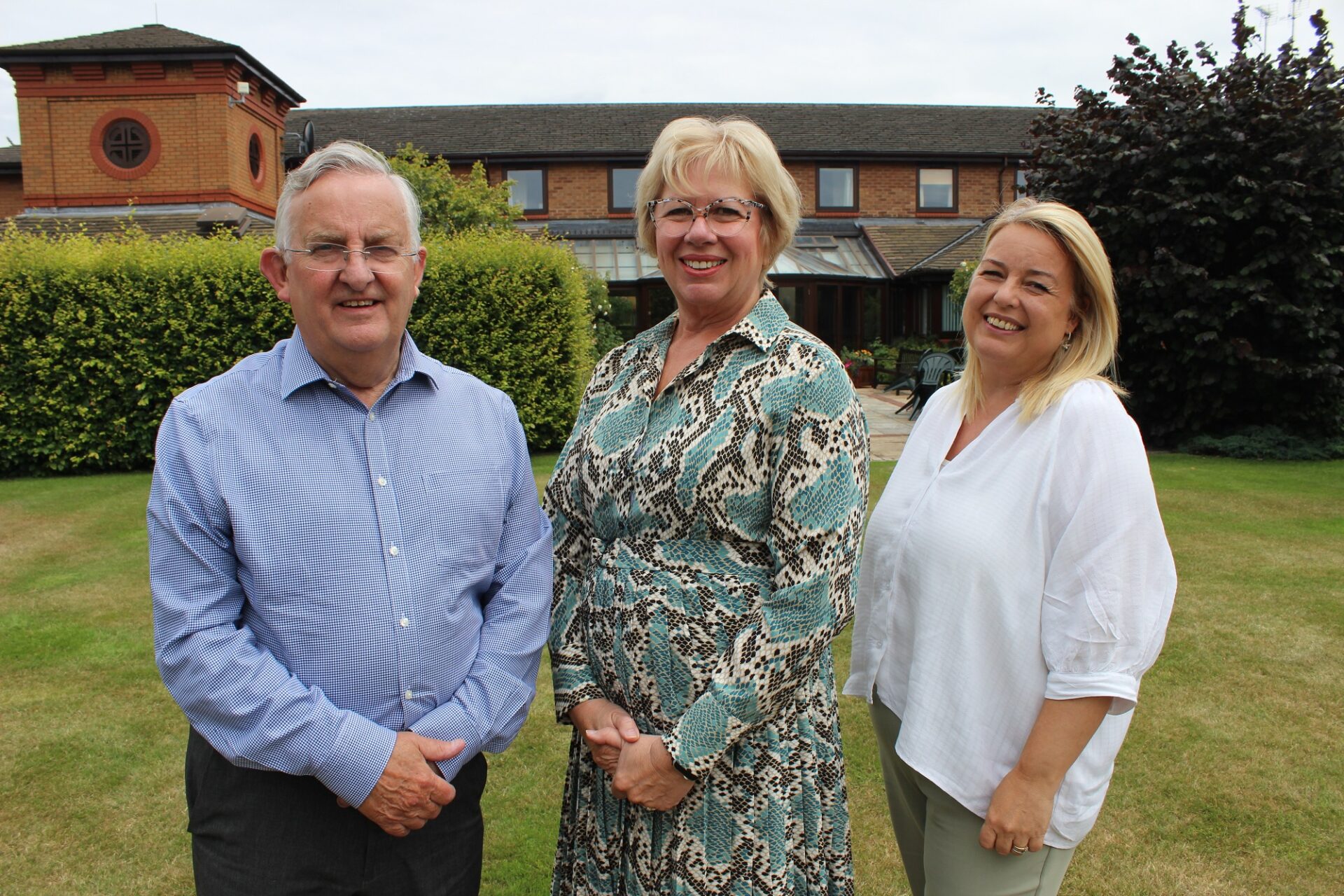 One man and two women stood on a lawn in front of a brick building