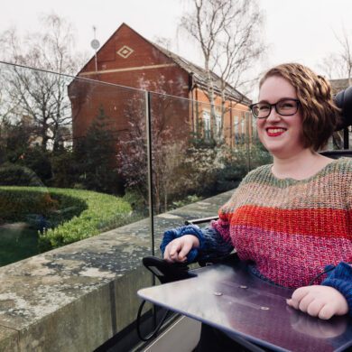Woman wearing a brightly coloured jumper sat in a wheelchair on a terrace smiling to the camera