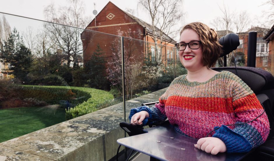 Woman wearing a brightly coloured jumper sat in a wheelchair on a terrace smiling to the camera