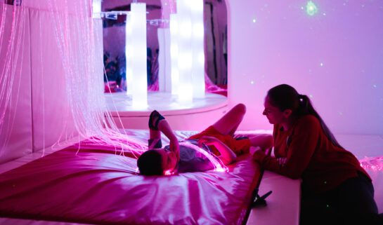 Child laying down in a sensory room filled with purple and coloured lights with a woman sat on the floor next to them.