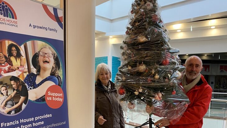 Man holding an artificial Christmas tree stood at the entrance to a shop door way next to a woman