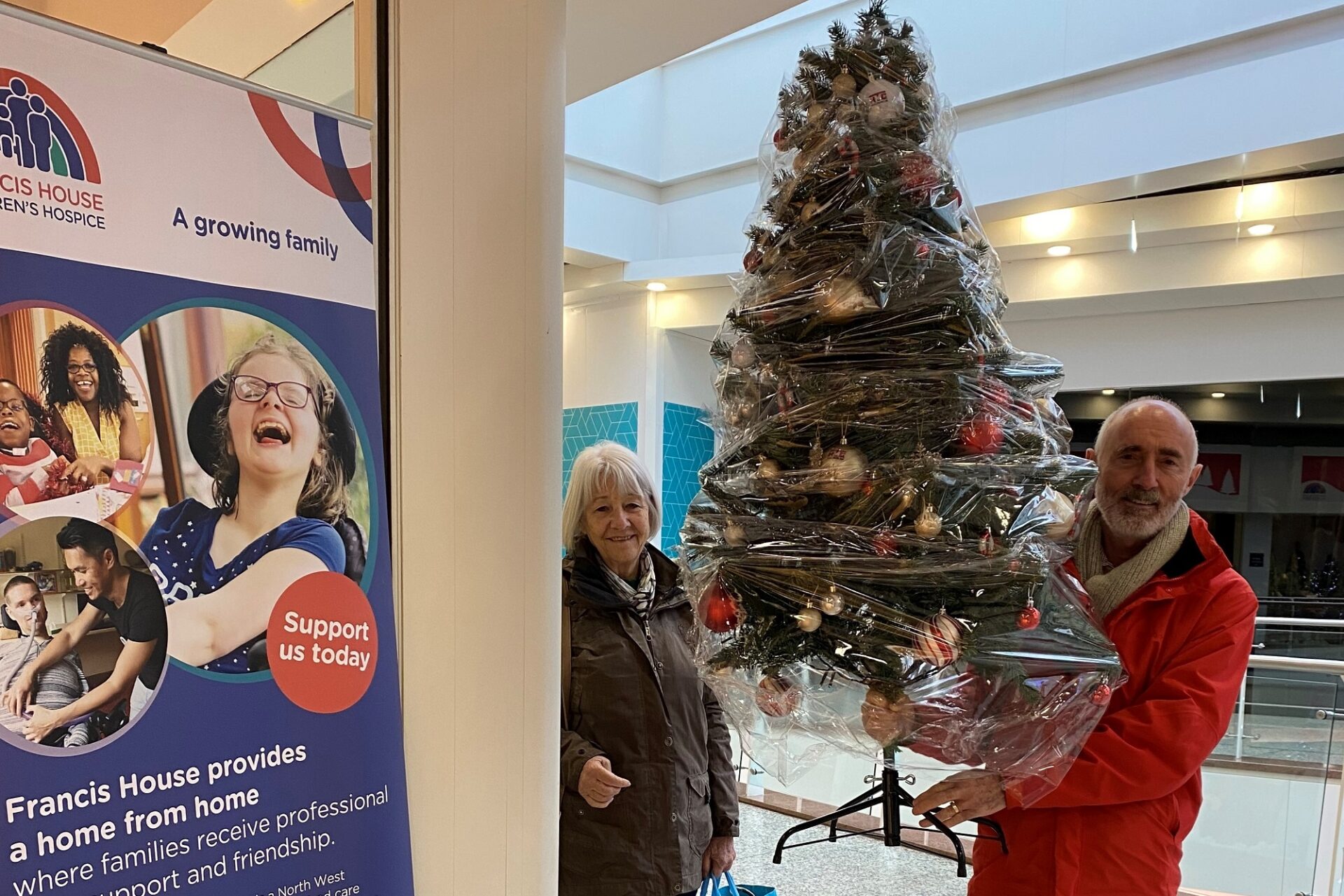 Man holding an artificial Christmas tree stood at the entrance to a shop door way next to a woman