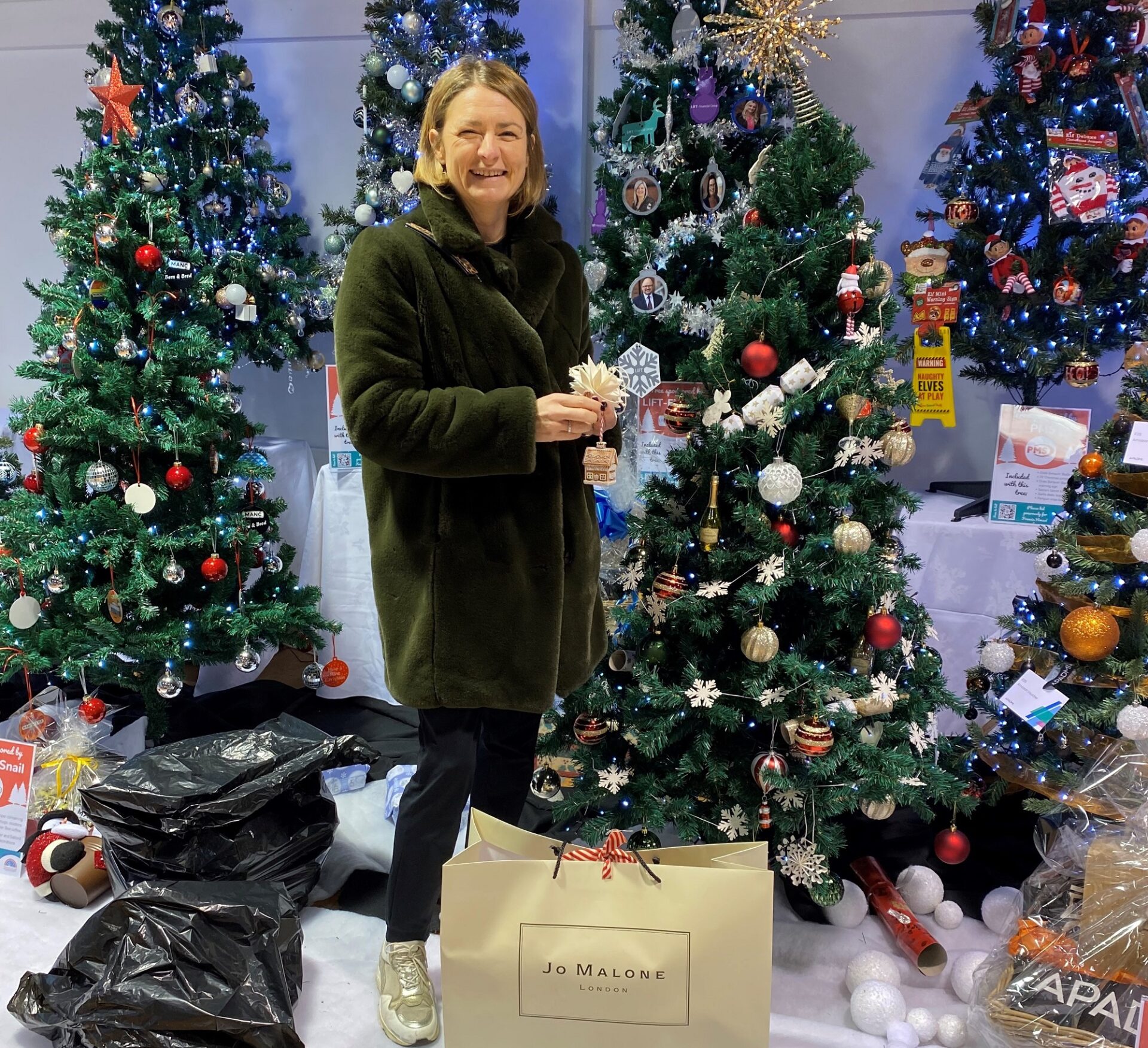 Woman wearing black coat holding a bauble stood in front of a Christmas tree with a large bag with the words Jo Malone on it