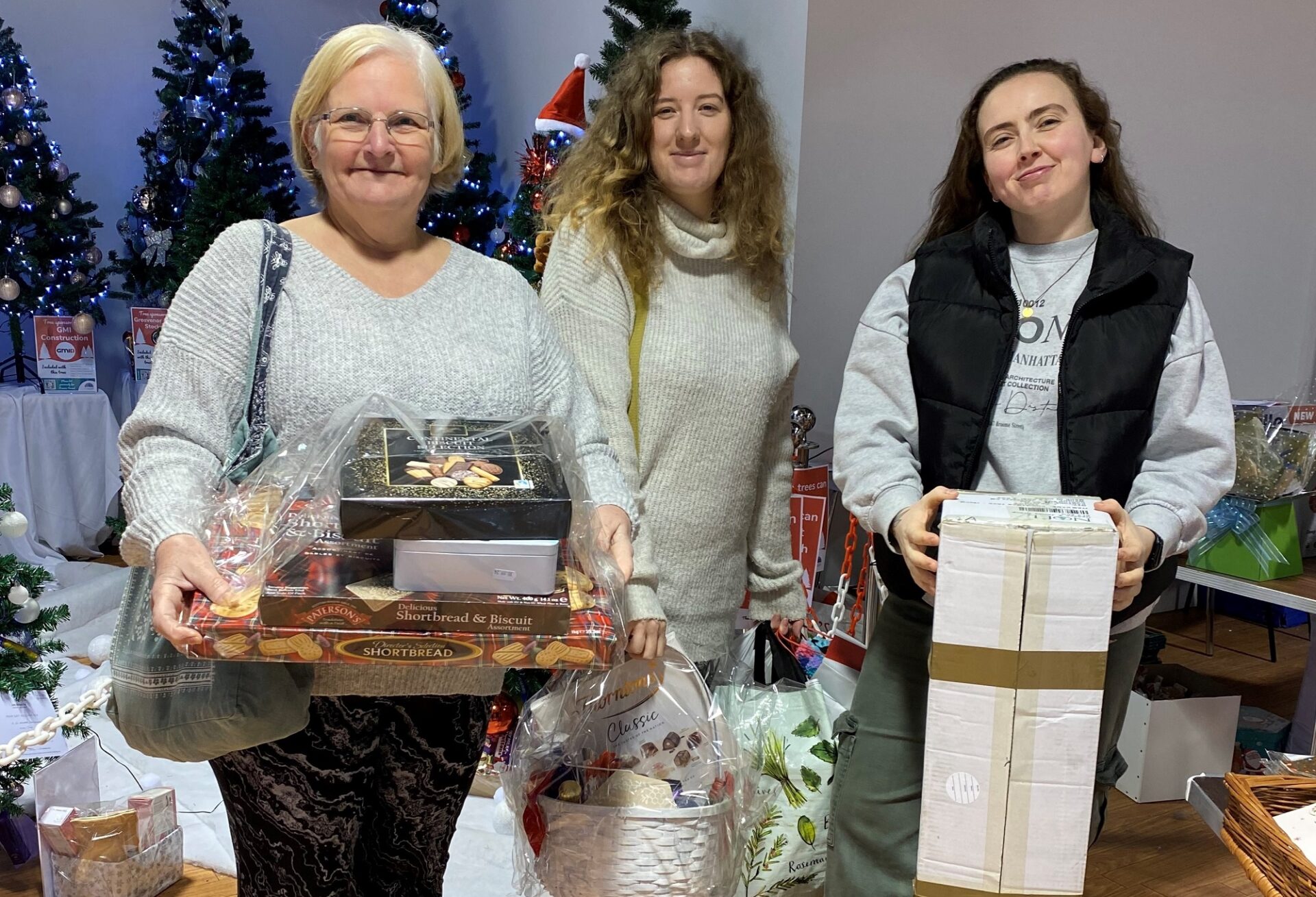Three women stood holding hampers and boxes in front of Christmas trees