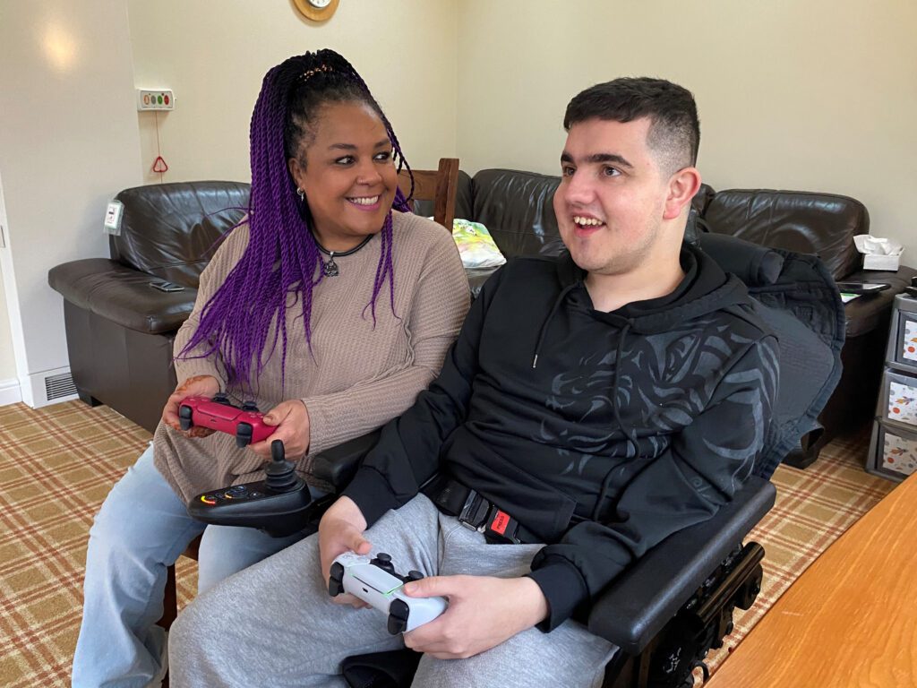 Woman holding a game keypad sat next to a young person sat in a wheelchair also holding a game keypad