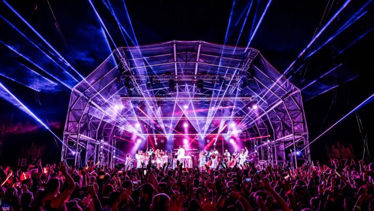 Orchestra on a stage lit by lights and lasers at night with a crowd of people stood in front of the stage