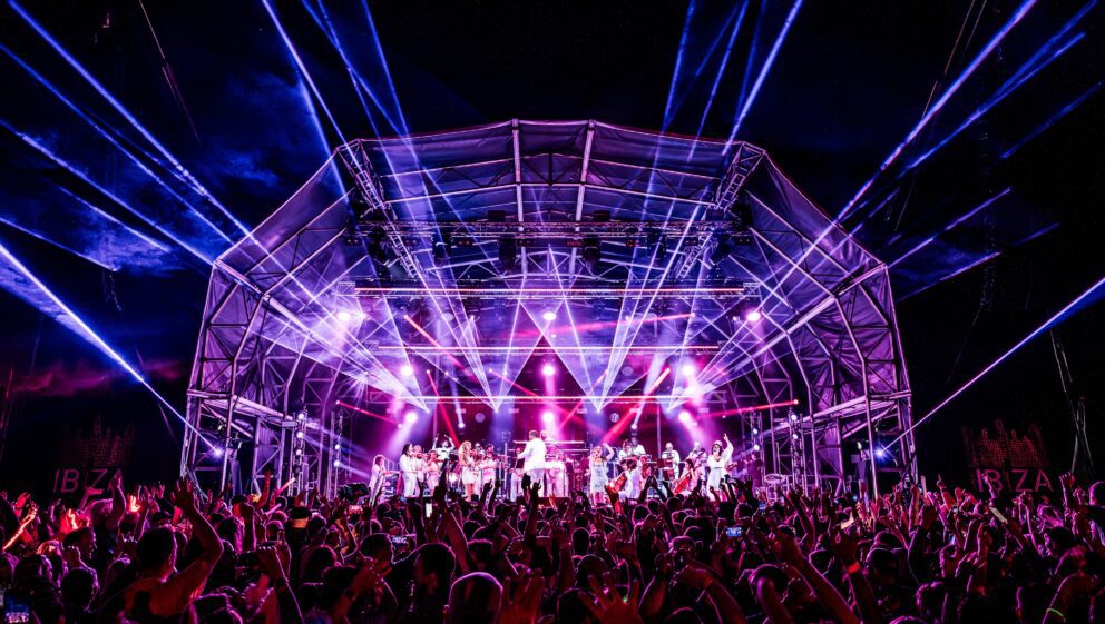 Orchestra on a stage lit by lights and lasers at night with a crowd of people stood in front of the stage