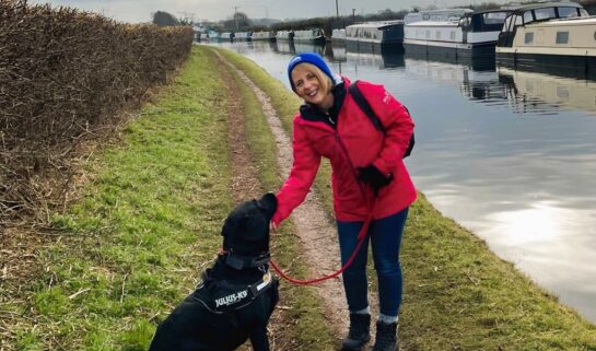 Woman wearing a red jacket and blue hat bending down to stroke a black dog on the tow path by a canal.