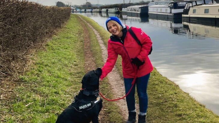 Woman wearing a red jacket and blue hat bending down to stroke a black dog on the tow path by a canal.