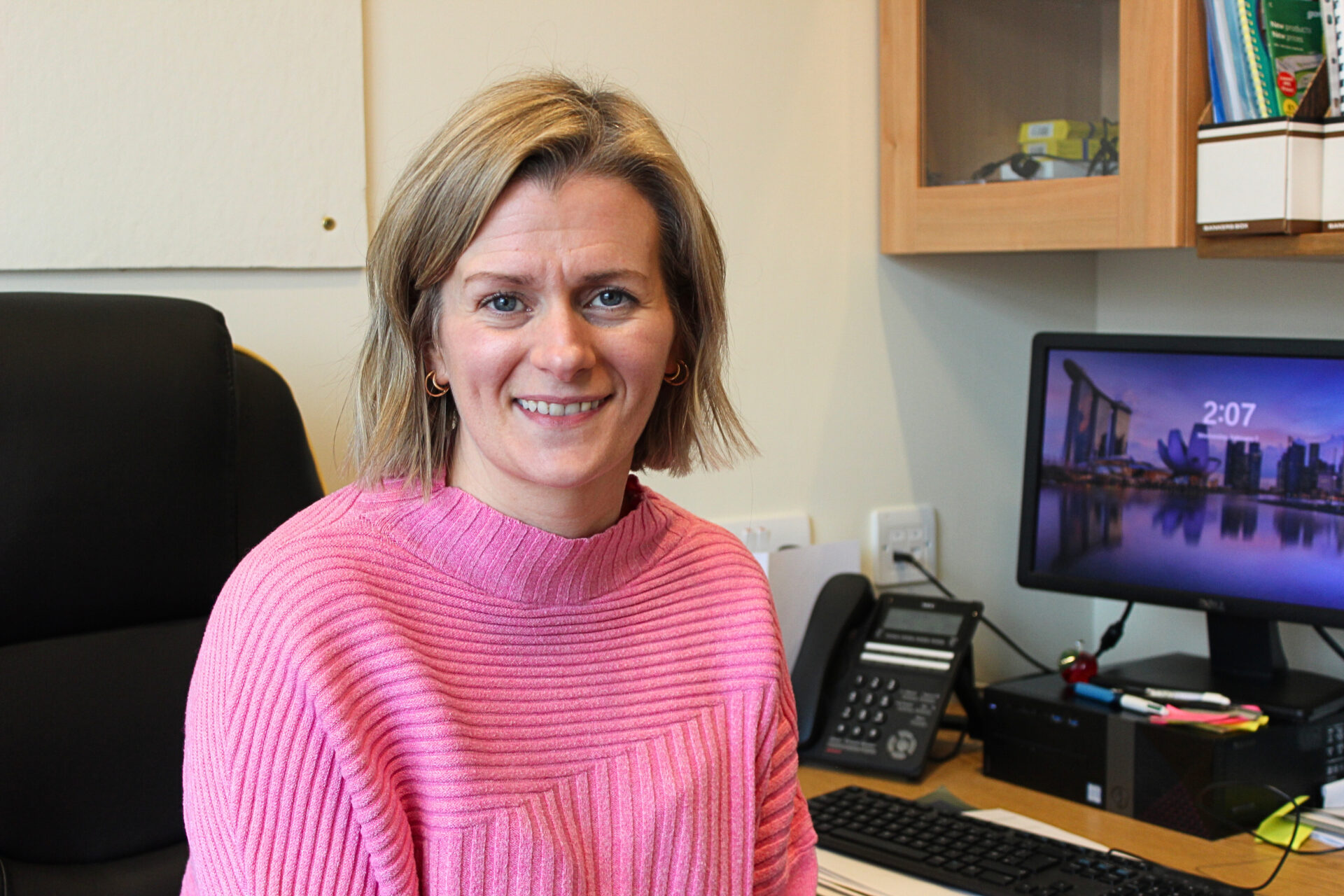 Woman wearing a pink jumper sat beside a computer in an office setting