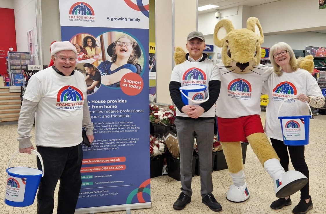 Three people wearing Francis House t-shirts stood holding buckets in a supermarket next to a large mouse mascot