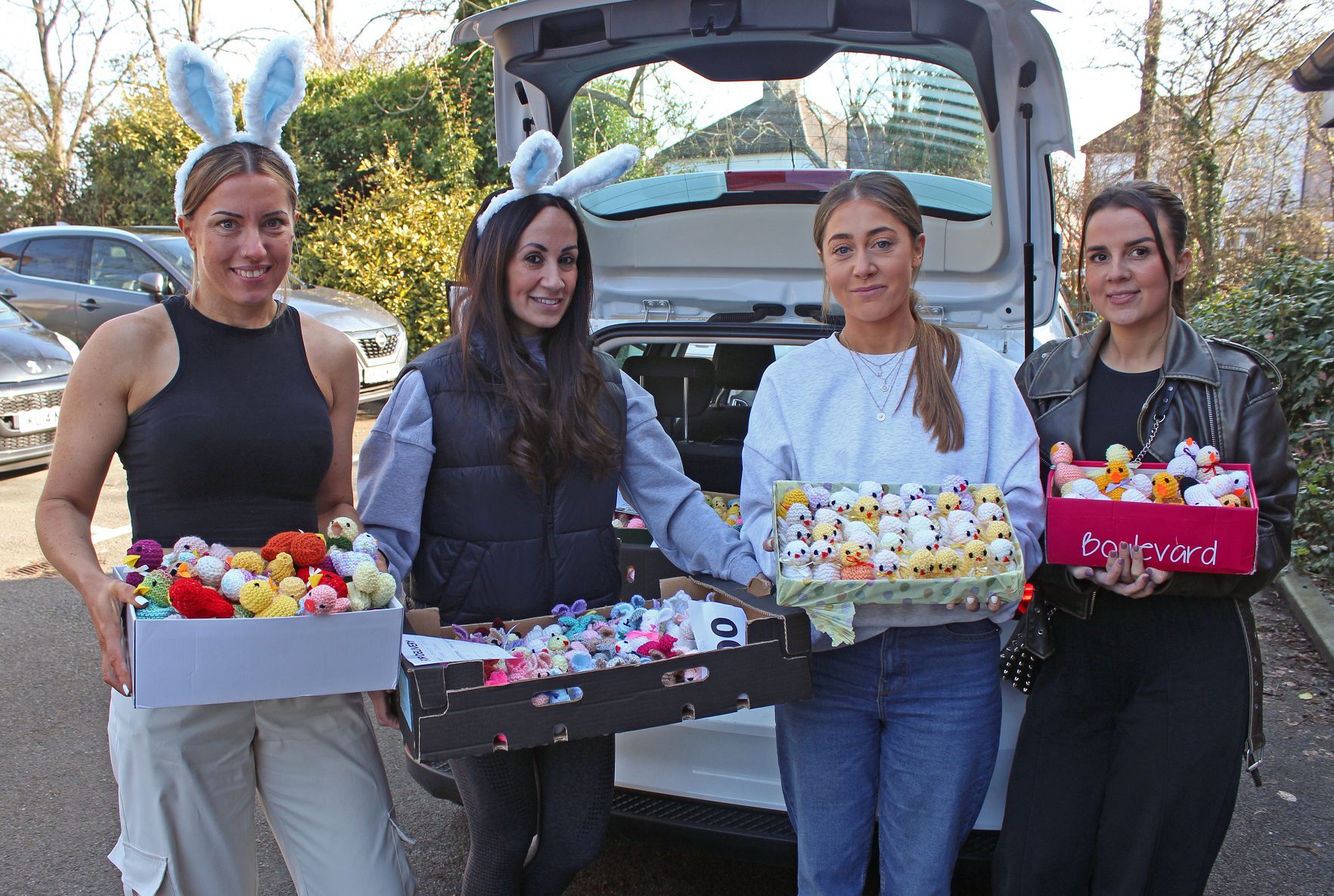 Four women holding boxes of knitted chicks stood in front of a white car with the boot open