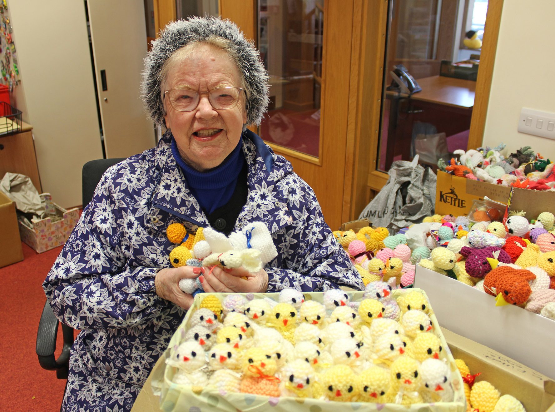 Woman wearing a woolly hat and patterned coat holding yellow knitted chicks sat behind a box of knitted chicks on a table
